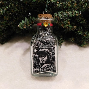 May include: A glass bottle ornament filled with black and white paper scraps with the words "BOB MARLEY" printed on the bottle. A black and white image of Bob Marley is visible through the paper scraps.