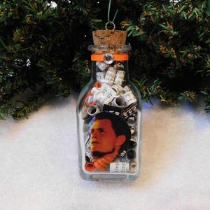 May include: A glass bottle ornament filled with rolled up pieces of paper with text printed on them. The bottle has an orange ribbon around the top and a small round sticker with a man's face on it.