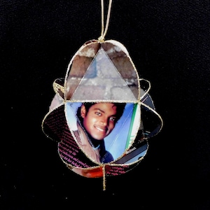 May include: A handmade ornament featuring a portrait of Michael Jackson. The ornament is constructed from layered paper and has a gold string for hanging. The design includes a triangular window and a circular shape.