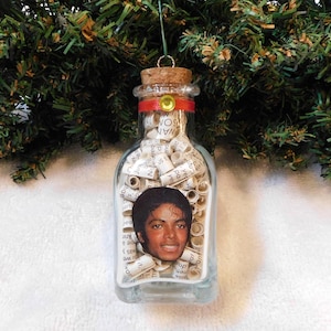 May include: A clear glass bottle ornament filled with rolled paper and a photo of Michael Jackson. The bottle has a cork top with a red ribbon and gold accents, hanging from a green string.