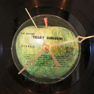 May include: A black vinyl record clock featuring the album art from The Beatles' "Yellow Submarine." The clock face displays a green apple graphic with gold-colored hands. The text "THE BEATLES YELLOW SUBMARINE STEREO" is visible.