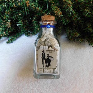May include: A clear glass bottle ornament with a cork stopper, filled with rolled paper scrolls. A blue ribbon with a small blue bead encircles the neck. A square label on the front features a black and white illustration and the text Fleetwood Mac Rumours.