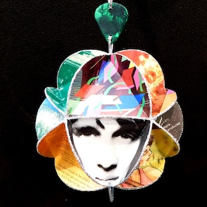 May include: A colorful, multi-layered paper ornament with a silver trim. The ornament features a face in the center and text that reads "You don't have to be afraid, Wherever you go you come back, I don't have to be..."