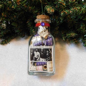 May include: A clear glass bottle ornament filled with rolled paper and a black and white photo of a band. The bottle is topped with a cork and red ribbon, and a purple gem. The ornament hangs from a green string.