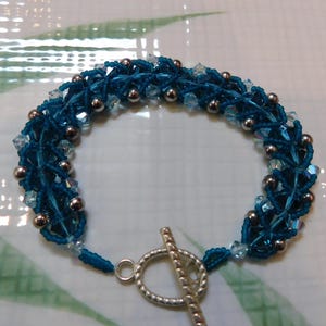 May include: A handcrafted teal bracelet with a woven design. The bracelet features small silver beads and clear crystal accents. It has a silver toggle clasp. The bracelet is approximately 7 inches in length.