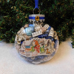 May include: A clear glass Christmas ornament filled with rolled up paper scraps and a paper cutout of a map with the words "CROSBY STILLS NASH YOUNG" printed on it. The ornament is decorated with a blue ribbon and a gold bead.