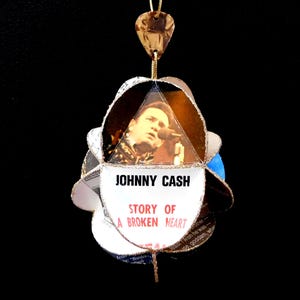 May include: A handmade ornament featuring a picture of Johnny Cash. The ornament has a multi-layered design with gold trim and a gold hanging cord. The text "JOHNNY CASH STORY OF A BROKEN HEART" is visible on the ornament.