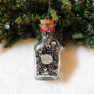 May include: A clear glass bottle filled with tightly rolled black and white paper strips featuring printed text. The bottle is sealed with a cork stopper, features a red ribbon with a purple gem, and a circular label reading Kenny Rogers.