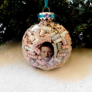May include: Clear glass Christmas ornament filled with rolled paper and a photo of Lionel Richie. The ornament has a silver cap with a teal ribbon and a purple gem. The ornament is hanging in front of a green Christmas tree.