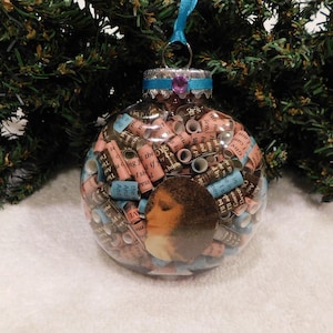 May include: A clear glass Christmas ornament filled with rolled up pieces of paper with gold lettering. The ornament is decorated with a pink and blue ribbon and a silver cap.