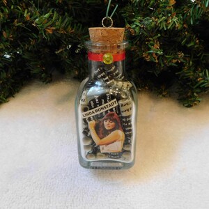 May include: A clear glass bottle ornament with a cork top and a red ribbon. The bottle is filled with rolled up pieces of paper with song titles and lyrics from Linda Ronstadt. The bottle has a label that says "Linda Ronstadt".