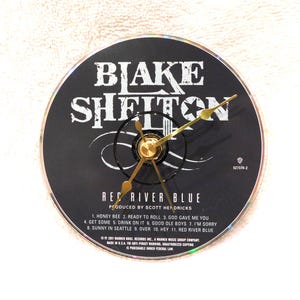 May include: A black clock with gold hands and a gold center. The clock face is a CD with the text "Blake Shelton" and "Red River Blue". The CD also lists song titles and production credits.