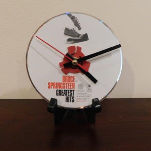 May include: A circular wall clock featuring a Bruce Springsteen Greatest Hits CD design. The white face displays a black and white image of shoes, red and black text, and a red plastic center adapter. It has thin red and black clock hands.