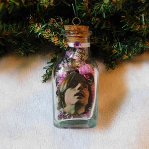 May include: A clear glass bottle ornament with a cork stopper, filled with small pink and purple paper rolls and a photo of a person. The bottle is decorated with a white ribbon and a purple gem. The text "You Stay" is visible.