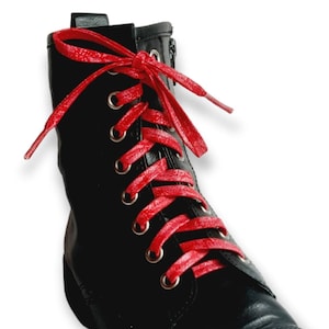 May include: Black leather combat boot with bright red laces. The boot features a rounded toe, a side zipper, and gold-colored eyelets. The laces are tied in a classic bow. The boot is set against a white background.
