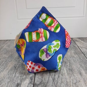 May include: A fabric bowl with a blue background featuring a repeating pattern of colorful flip-flops in various designs, including green stripes, red polka dots, and floral prints. The bowl is secured at the top corners with a white plastic snap fastener.