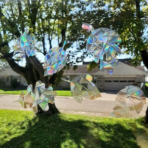 May include: Seven iridescent holographic leaf stickers, each with a unique geometric pattern, are displayed on a window. The stickers are a light blue, silver, and gold color.