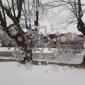 May include: Iridescent snowflake window clings in various sizes and shapes, creating a winter scene. The clings are applied to a window, with a snowy outdoor backdrop featuring bare trees and houses.