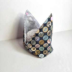 May include: A fabric yarn bowl with a dark blue background covered in various colorful buttons. The interior is a light gray patterned fabric. A ball of gray yarn sits inside the bowl, held in place by pink snap closures.