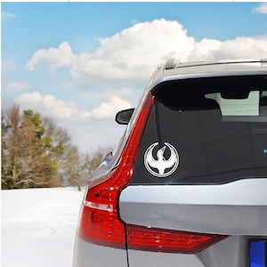 May include: A white vinyl decal of a stylized bird, possibly a phoenix, is affixed to the rear window of a silver SUV. The bird is encircled by a simple border. The vehicle's taillight is red, and the background shows a snowy landscape and a blue sky.