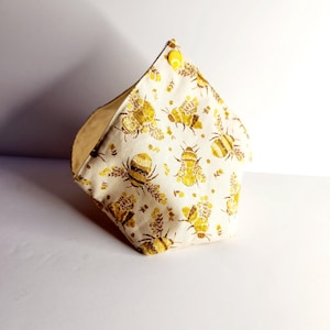May include: A fabric container with a bee pattern. The container is made of a cream-colored fabric with a repeating pattern of yellow bees and floral elements. A yellow button secures the top.