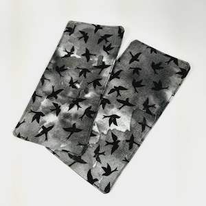 May include: Two rectangular fabric items with a black bird silhouette pattern on a gray and white background. The edges are neatly stitched. The design features a flock of birds in flight, creating a dynamic and airy feel. The items appear to be decorative or functional.