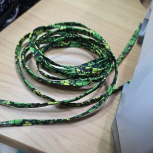 May include: Coiled green and black patterned shoelaces. The laces have a repeating design of yellow and black shapes on a green background. The laces are on a light brown wooden surface.
