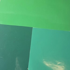 May include: An overhead shot of four sheets of paper in shades of green. The sheets are arranged in a geometric pattern, with the colors ranging from light to dark green. The paper has a glossy finish.