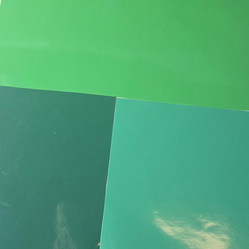 May include: An overhead shot of four sheets of paper in shades of green. The sheets are arranged in a geometric pattern, with the colors ranging from light to dark green. The paper has a glossy finish.