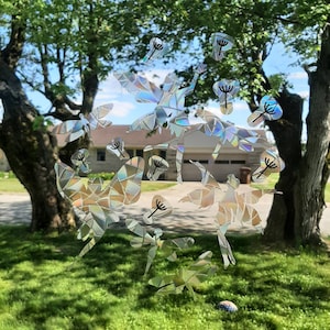 May include: A window cling featuring a design of iridescent dragonflies and dandelion seed heads. The shapes are made of geometric shapes that reflect light. The cling is placed on a window with a view of a house and trees.
