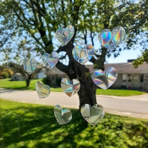 May include: A window with a view of a green lawn and a street. There are 10 iridescent heart-shaped stickers on the window. The stickers are made of a clear material with a rainbow-like finish.