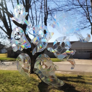May include: Seven iridescent ghost window clings. Each ghost has a unique shape and design, with a mosaic-like pattern that reflects light. The ghosts are in various poses, creating a playful and spooky Halloween decoration.