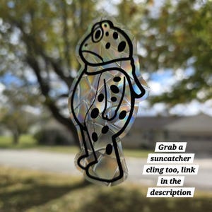 May include: A frog-shaped suncatcher cling with a textured, iridescent surface and black outline. The frog has black spots and a scarf. Text on the image reads "Grab a suncatcher cling too, link in the description."
