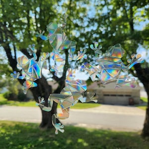 May include: Five iridescent frog silhouettes are clustered together on a window. The frogs are made of a reflective material that creates a rainbow effect.