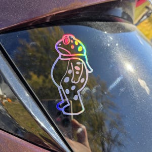 May include: Holographic sticker of a cartoon frog with a sad expression. The frog has a round body, a small head, and is wearing a scarf. The sticker has a rainbow sheen and is applied to a dark surface.