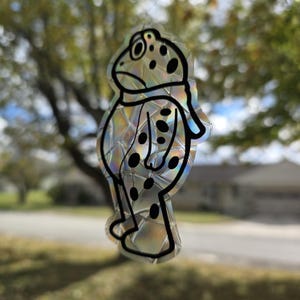 May include: A clear, iridescent frog-shaped window cling with black outlines and black spots. The frog has a scarf and is standing upright. The background is blurred, showing a street and trees.
