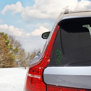 May include: A green vinyl car decal featuring a cartoon frog wearing a scarf. The decal is applied to the rear window of a silver car. The background shows a snowy landscape and a blue sky with clouds.