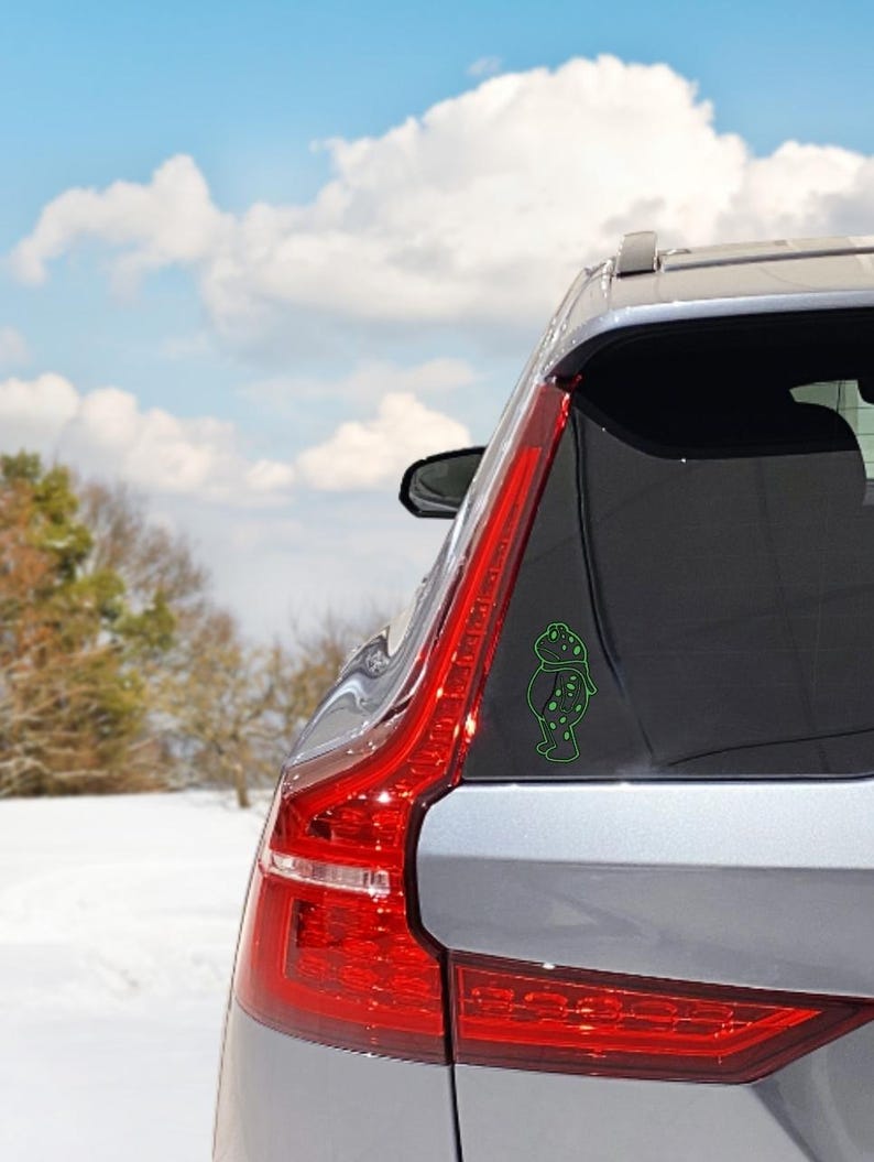 May include: A green vinyl car decal featuring a cartoon frog wearing a scarf. The decal is applied to the rear window of a silver car. The background shows a snowy landscape and a blue sky with clouds.