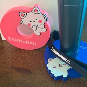 May include: A pink circular stand with the text SAKKUKK and a white cat illustration, alongside a blue circular stand featuring a white cat figure and a black strap with a white graphic design, supporting a dark gray cylindrical device.