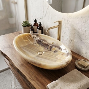 Puede incluir: Un mueble de baño con encimera de madera natural y un lavabo ovalado con efecto mármol. El lavabo tiene una combinación de colores amarillo claro y marrón. También se ven un grifo dorado, un dispensador de jabón y una pequeña planta suculenta.