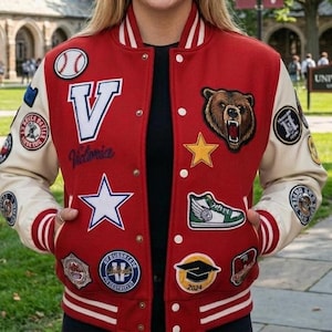 May include: Red varsity jacket with cream sleeves and various embroidered patches. Includes a large white "V" with "Victoria" script, a bear, star, baseball, and a graduation cap. The jacket has white stripes on the cuffs and hem.