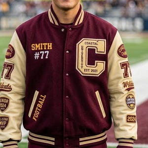 Może przedstawiać: Bordowa i kremowa kurtka uniwersytecka z napisem "SMITH #77" i "COASTAL UNIVERSITY EST. 2021". Kurtka posiada wiele naszywek, w tym "FOOTBALL", "Wildcats" i "2024".