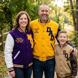 May include: Three varsity jackets featuring embroidered text. The purple jacket has white sleeves and says MOTHER OF THE YEAR EST. 2012 and MOM. The yellow jacket has black sleeves and says FATHER THE LEGEND and DAD. The tan jacket has brown sleeves and says THE GOAT TRAINING EST. 2016 and SON.