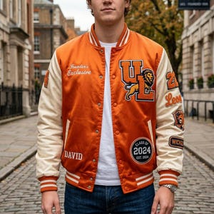 Könnte beinhalten: Orangefarbene Satin-Collegejacke mit cremefarbenen Ärmeln. Mit dem Text "Founder's Exclusive" und "DAVID". Enthält Aufnäher mit einem Löwen, den Buchstaben "UL" und "Class of 2024".