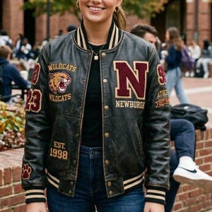 Puede incluir: Chaqueta universitaria de cuero negro con ribete a rayas doradas y blancas. La chaqueta presenta parches con las palabras "Wildcats", "Newburgh" y "EST. 1998". Una gran "N" está en el pecho.