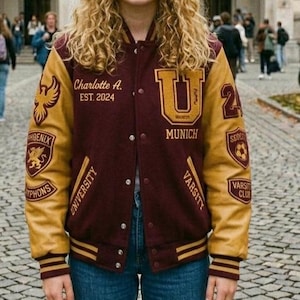 Pode incluir: Casaco universitário bordô e dourado com o texto "Charlotte A. EST. 2024" e "Munich" na frente. O casaco tem patches com uma fénix e outros emblemas. As mangas são douradas e o corpo bordô.