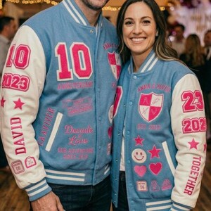 Può includere: Giacche varsity azzurre e bianche con dettagli rosa. Una giacca presenta "10" e "2023" sul davanti e "DAVID" sulla manica. L'altra giacca ha "23" e "2023" sulla manica e "ANNA & DAVID" sul davanti. Entrambe le giacche hanno varie toppe.