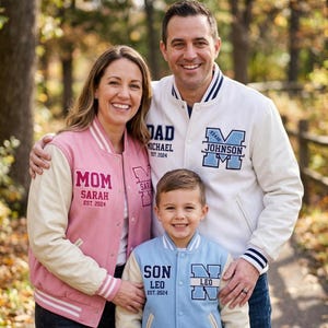 Peut inclure: Trois vestes style universitaire avec boutons-pression et bords-côtes rayés. La veste rose porte l'inscription 'MOM SARAH EST. 2024', la blanche 'DAD MICHAEL EST. 2024' et 'JOHNSON', et la bleu clair 'SON LEO EST. 2024'.