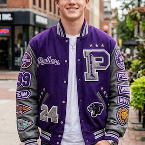 Puede incluir: Chaqueta universitaria morada y gris con la palabra "Panthers" y una gran letra "P". La chaqueta tiene varios parches, incluyendo "STATE CHAMPIONS 23", "99 TEAM" y un logotipo de pantera. Las mangas son grises con rayas blancas.