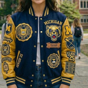 Op de afbeelding: Een marineblauwe en gouden varsity jas met meerdere patches. De jas heeft het woord "MICHIGAN" en een berenafbeelding. Patches zijn onder meer "UNIVERSITY OF MICHIGAN 1817", "SARAH", "SENIOR '25" en "HONORS COLLEGE".
